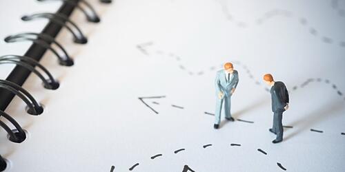 image of two miniture plastic men in suits looking down at penned dotted arrows on paper